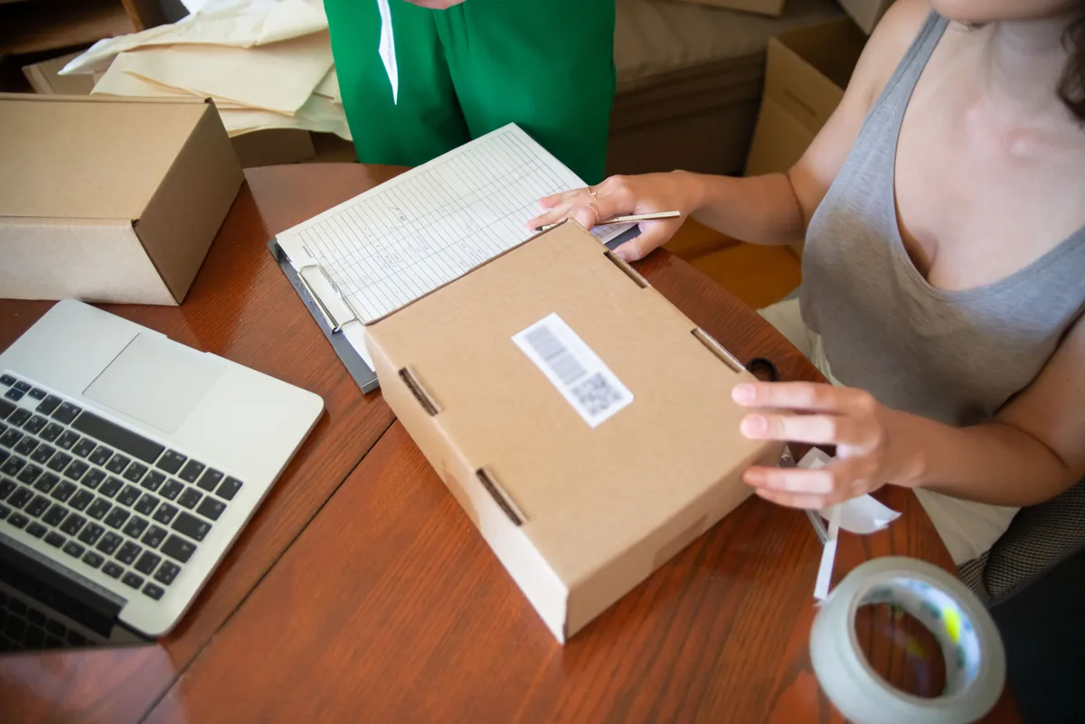 A person checks a parcel beside a laptop and order sheet on a wooden work table.