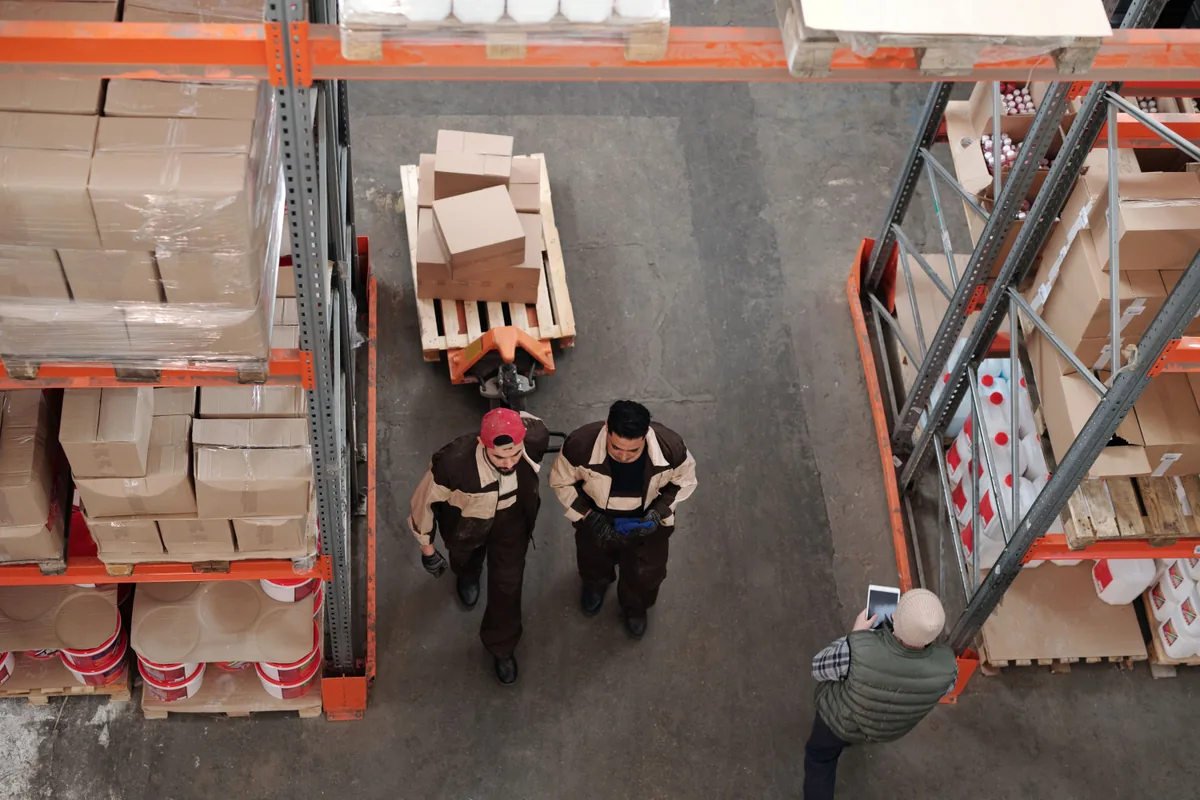 Overhead view of cartons on pallets, a pallet truck and workers in a warehouse aisle.